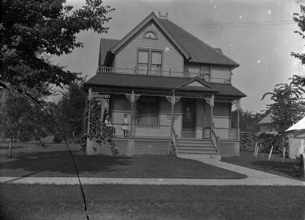 View from street towards a man and a girl posing on the porch of a two-story wood-frame house. Identified as the Ed Locken Residence, northeast corner of 4th Street and Van Buren. Fuelwood is stacked behind the house on the right.
