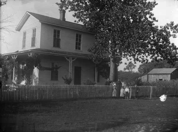 View towards a man, a woman and four children standing posing near the picket fence of a two-story wood-frame house. Identified as Residence built by A. Hyslop, and inhabited by Johnny Johnson. There is a barn in the background on the right. Located at the southwest corner of South 2nd Street and Johnson Street.