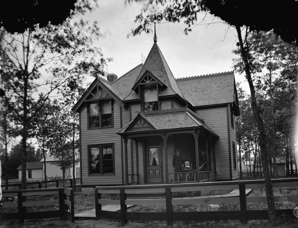 View across road and fence towards a two-story wood-frame house. A man is standing on the porch next to an infant in a baby carriage. A wooden sidewalk leads to the front porch, and also curves around the side of the house on the right. Identified as the residence of Petra Kjorstad at 417 Monroe Street. There are buildings in the background on the left and right.
