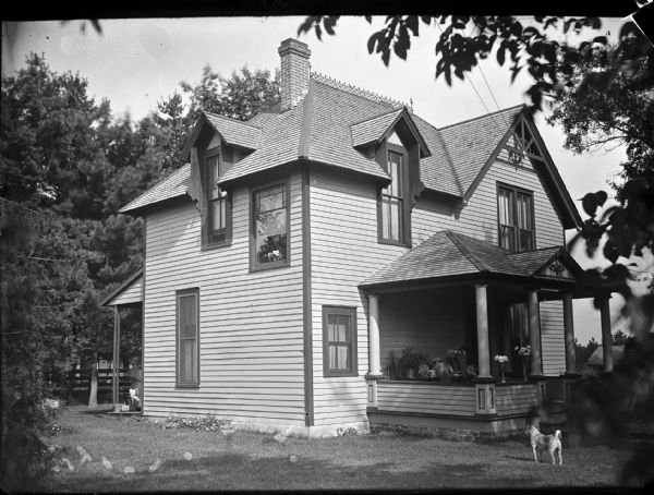 Outdoor view across lawn towards a two-story wood-frame house. Flowers in vases and plants in pots are arranged on the porch, and a dog is standing in the yard on the right. Just behind the left corner of the house which has a back porch, is an infant sitting in a baby carriage and wearing a bonnet. Identified as the residence of William O'Hearn, later Dr. Kalling. Located at the southwest corner of North 5th Street and Monroe Street.