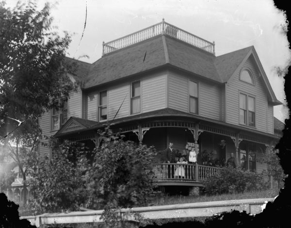 View across fence and lawn towards a two-story wood frame house with a wrap-around porch. A man, two women and two children are standing on the porch. Identified as the W. Maddocks Residence, 119 South 2nd Street.