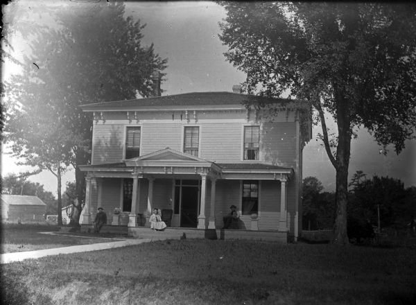 View across lawn towards the front of a two-story wood-frame house. One European-American woman and two European-American men wearing hats are sitting on the front porch. Pottery vessels are displayed on pedestals on the left and right side of the porch. The home is identified as the Roddy residence.