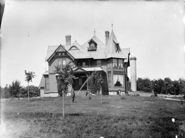 View across lawn towards a large wooden house with many gables. A girl is standing under a front window of the house on the right, and a man is standing in the center watering a young tree in the lawn with a hose. There are other small trees planted in the yard. Identified as the O'Hearn residence. Behind the house on the right is a tall tower.