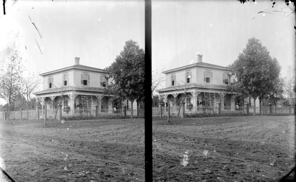 A stereoscopic outdoor view across unpaved road towards a two-story wooden house surrounded by fencing. Identified as the residence of J.J. McGillivray; northwest corner of Eighth and Main Streets. A group of people are posing on the porch, including a woman standing at the left corner of the porch, a group of four men and an infant in a baby carriage in the center at the corner of the porch, and a woman on the far right of the porch, standing in a doorway.