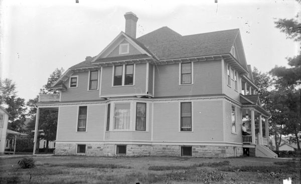 View across lawn towards the side of a two-story wooden house with a porch in the front on the right, and a balcony in the back on the left. A woman is sitting on the porch rail. The home is identified as the residence of R.P. Rainey, located at 6th and Pierce Streets, and was built in 1909.