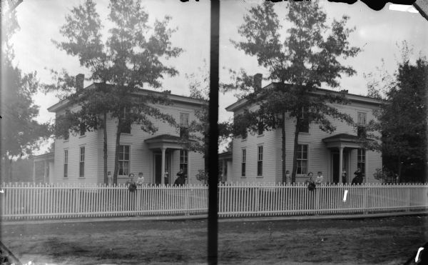 View across unpaved street towards a two-story wood frame house. Two woman and three children are posing, with a woman and child at the fence surrounding the yard in the foreground, a girl standing between two trees on the left, and a child standing and a woman sitting on the porch under the roofed entrance at the front door. Identified as the residence of W.T. Murray, 1008 Main Street.