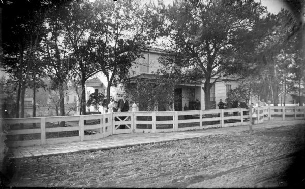 View across unpaved street towards a group of people posing in front of a two-story wooden house. A light-colored fence is along the length of the front yard, with man and a boy standing at the gate on the left. Two women are on the porch, and two women are standing between the windows. Two women and a girl are standing next to the fence. Identified as the Le Clair residence located at the southeast corner Main and Melrose Streets.