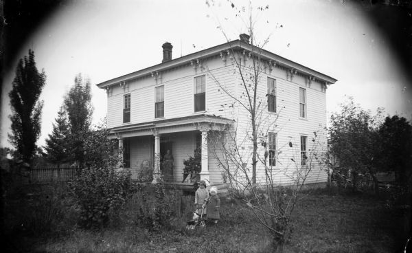 View across lawn towards a two-story wooden house. A woman is standing on the porch, and two children are standing in the yard. The older child is holding onto a baby carriage with a doll in it. Identified as the Tom Roddy residence located at 307 S. Second Street.