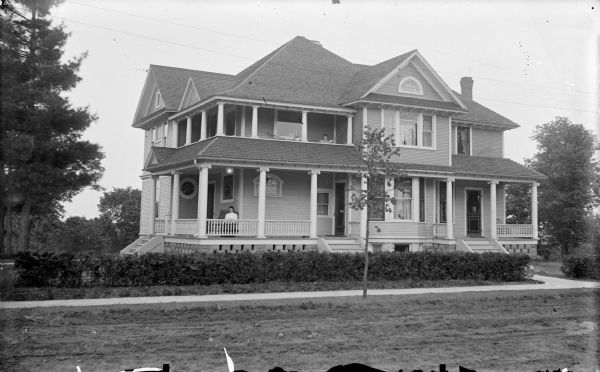 View across unpaved street towards a two-story wooden house with a wrap-around porch and three entrances. A woman is sitting on the porch, and another woman is sitting in a hammock on the balcony. A row of bushes is along the sidewalk. Identified as the R.P. Rainey residence located at the southeast corner of 6th Street and Pierce Street. The house was built in 1909.