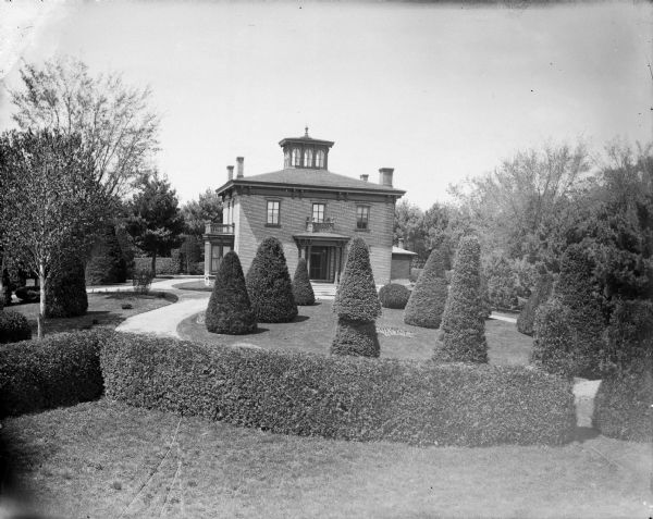 Slightly elevated view of a two-story brick house with a well-manicured yard with sculpted shrubs. A small balcony is in the front, and another is on the side. Identified as the Spaulding residence located at 1206 Harrison Street.