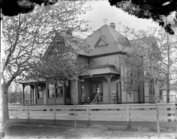 View from street towards an unidentified wooden house with a fence in front. A woman and three children are standing on the landing by a side entrance. Ironwork is on some of the gables.