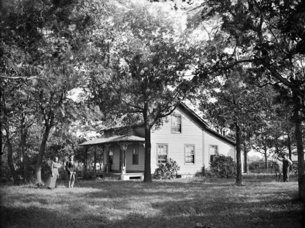 View across lawn towards an unidentified wooden house surrounded by trees. A man and woman are standing in the yard on the left, and a man and a horse are standing in the yard on the right.
