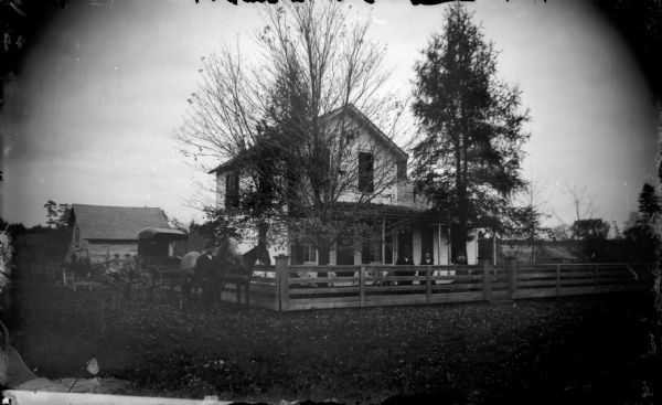 View from unpaved street towards a wooden house surrounded by a wooden fence. A man is standing next to two horses pulling a buggy which is parked outside of the fence on the left. A man and woman are standing inside the fence, and a woman is standing next to a tree in the yard on the right. A barn or stable is in the background on the left.