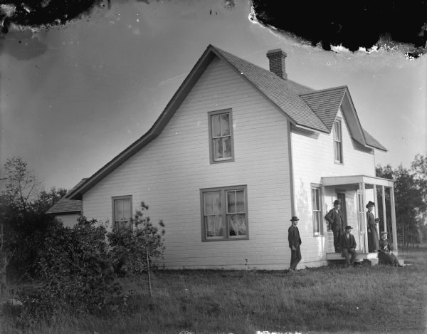 View towards an unidentified two-story wooden house with a group of three men and two woman posing on or near the front landing.