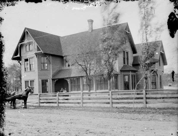 View across unpaved road towards a two-story wooden house with a screened-in porch. A horse and buggy are parked near the fence on the left. A woman and two children are sitting on the front steps.