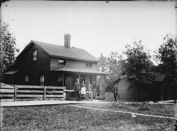 View towards a group of people in front of a small two-story wooden house. Three women are standing on the porch, and a man is leaning against the fence. A man in uniform is posing on the right with a sword.