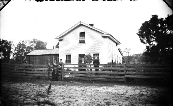 View towards a two-story wooden house. Three women and a boy are standing at the fence in front. A wooden structure is on the left.