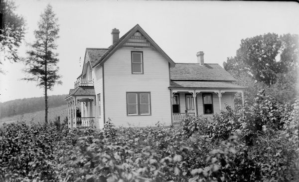 View across yard full of plants towards a two-story wooden house with roofed porch entrances on the side and in front, a balcony, and gable decorations and porch brackets. A field on a hill is in the background.