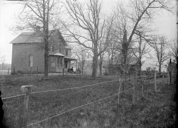 View across fence towards a two-story brick house. A man and two children are sitting on the porch. Two farm buildings are in the background on the right.