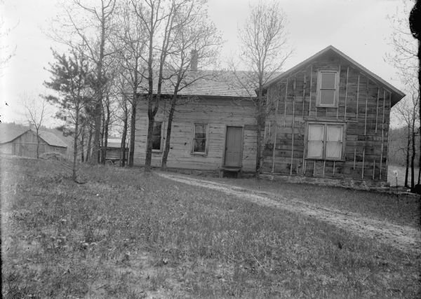 View towards a small two-story wooden house. Farm buildings are in the background.