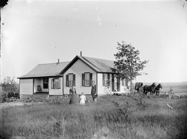 Outdoor group portrait of a man, woman and child posing in the tall grass in front of a small one-story wooden house with a small porch. A man is sitting in a buggy drawn by two horses on the right next to the house. In the distance are fields and hills.