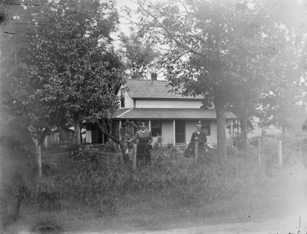 View towards a man and woman standing behind a fence in the yard of a small two-story wooden house.
