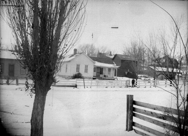 View across snow-covered ground towards two wooden houses with fences in front. A man is standing at the gate.