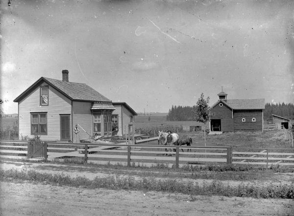 View across unpaved road and fence towards a group of people posing in front of a two-story wooden dwelling on the left, and a stable. on the right. A man and two horses are standing in the yard in the center. A woman is standing at the corner of the house, and two small children are sitting together in a hammock strung from the side of the house to a wooden post which is across a wooden sidewalk. A wood structure near the post may be a well. In the background is a buggy at the stable door.