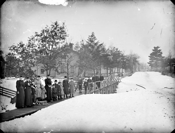 Exterior portrait of a group of women and children standing on a wooden sidewalk and bridge in a residential neighborhood on a snowy day.
