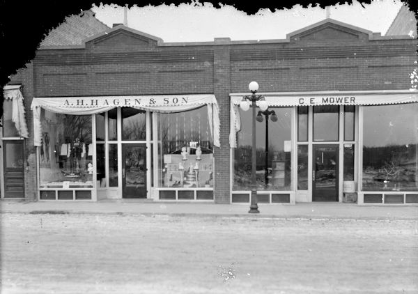 View across Main Street, between First and Water Streets. Businesses include A.H. Hagen & Son on the left, and C.E. Mower on the right. A lamppost is in the center.