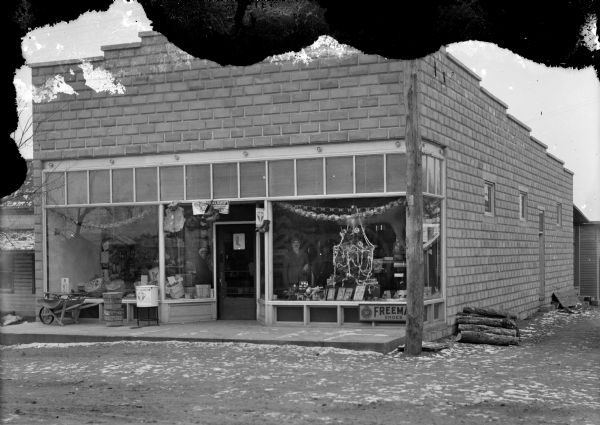 View across street towards the George Adams Store, located on the east side of South Roosevelt Road. A decorated Christmas tree is in the front window, and a woman and a man are standing inside the store looking out.