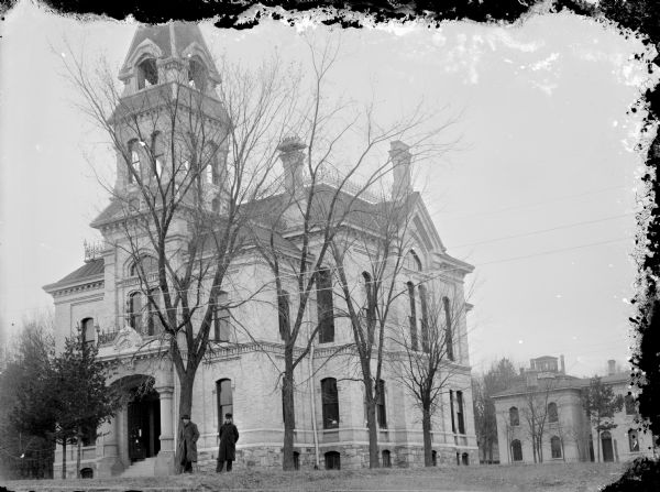 View of the Jackson County Courthouse. Two men are standing under one of the trees in front. Another building is in the background on the right.