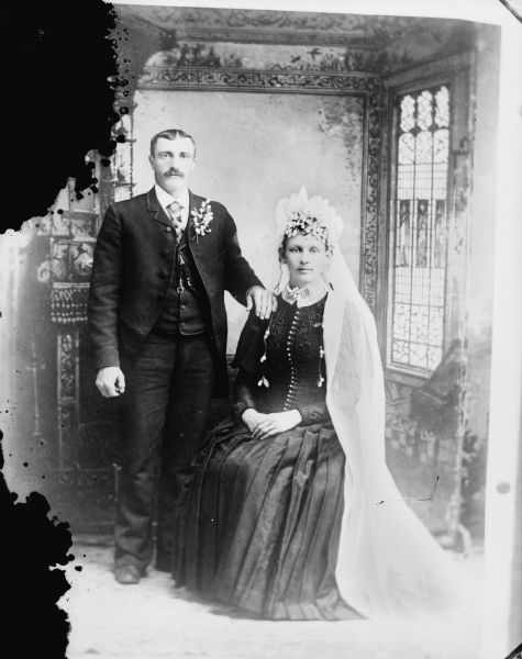 Studio portrait in front of a painted backdrop of a married couple. The man is wearing a suit and corsage. The woman is wearing a bridal veil and dark-colored dress.