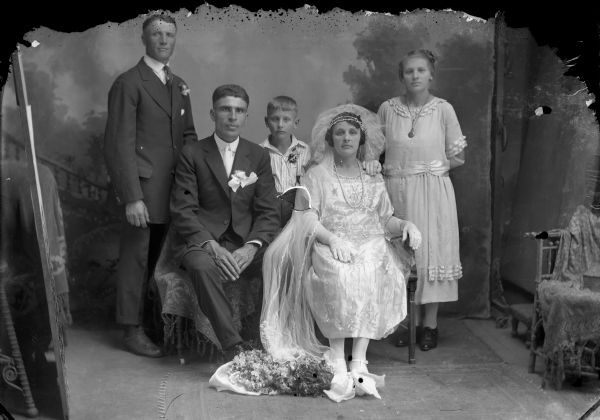 Studio group portrait in front of a painted backdrop of a wedding party, including two men, a woman, a girl and a boy. The bridal bouquet is at the bride's feet.