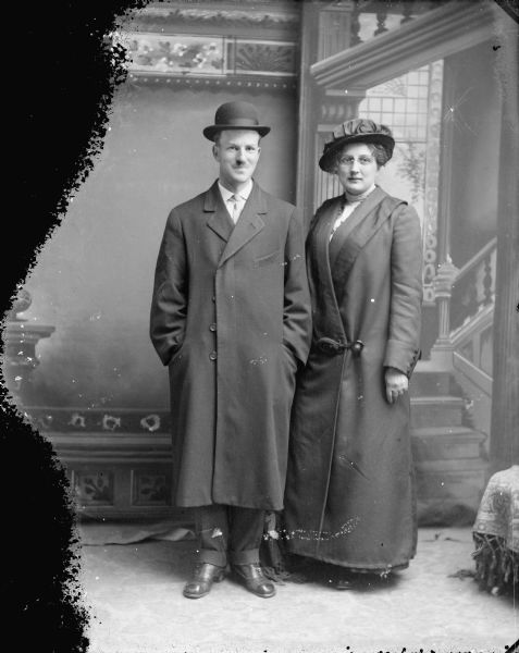 Studio portrait in front of a painted backdrop of a man and woman posing standing wearing coats and hats. The couple is identified as Mr. and Mrs. Roy Van Schaick.