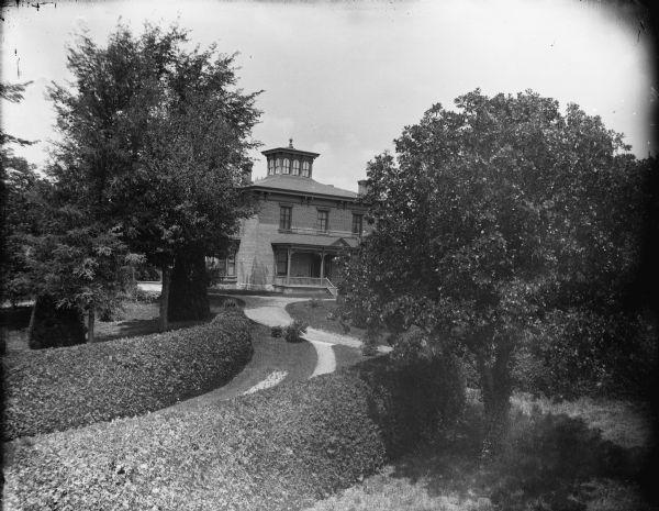 Elevated view across a well-manicured yard with sculpted shrubs, trees and front walk towards a two-story brick house, identified as the Spaulding residence.
