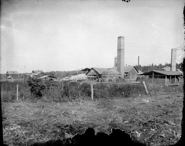 View of farm buildings and smokestacks beyond a fence in the foreground.