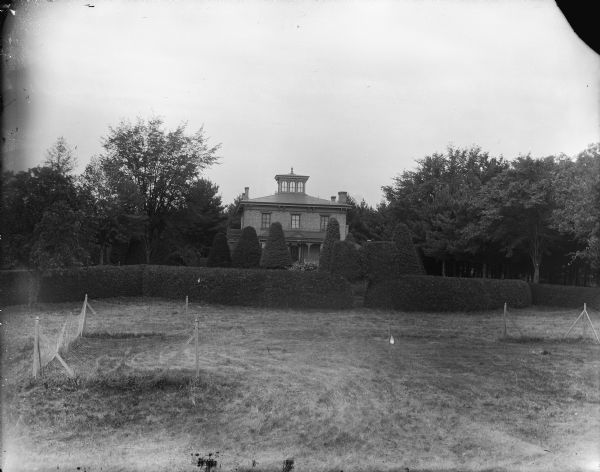 Elevated view across field with a two-story brick house and a large yard with manicured bushes and trimmed hedges. Identified as the Spaulding residence, with a well-manicured yard with sculpted shrubs.