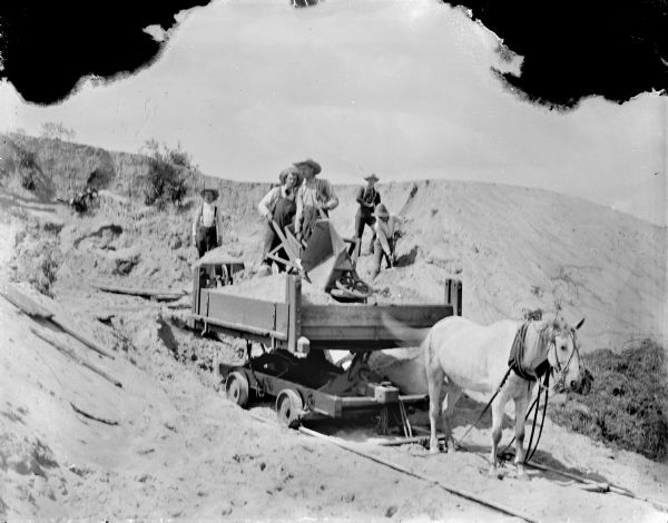 View of a horse hauling dirt on a wagon on tracks. There are five workmen adding dirt to the wagon from a large pile. Identified as part of the Hatfield Canal construction.