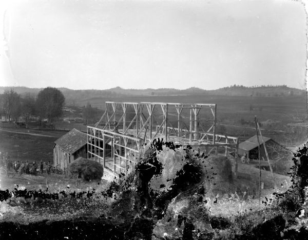 Elevated vis iew of a barn raising. One man standing in the framework, and a group of men are standing on the left.