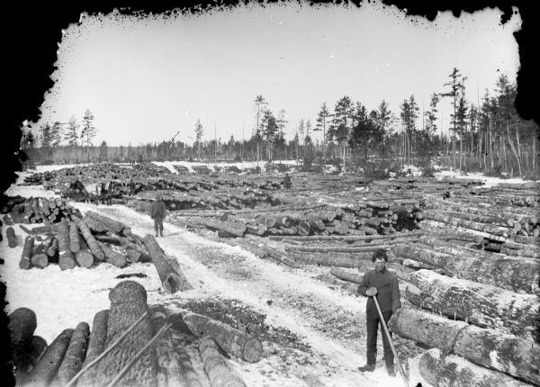 Elevated view of logs arranged in piles in a clearing with snow on the ground. Two workmen are standing on a pathway between them. In the background is a man standing near two horses pulling what may be a sled, which is stacked with logs.