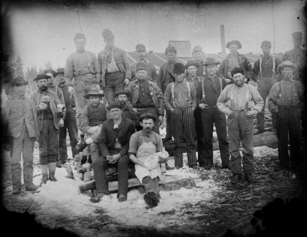 Outdoor group portrait of a group of workmen, probably at a lumber camp. The man sitting in the front in the center is holding a rabbit. A building is in the background.