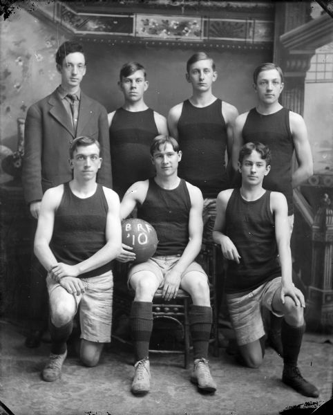 Studio group portrait in front of a painted backdrop of the 1910 Black River Falls basketball team. Three men are posing sitting, and four men are posing standing.