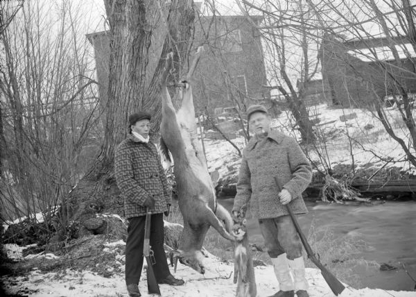 Exterior portrait of two hunters posing with a deer carcass, a fox carcass and rifles on the edge of a stream in winter. Buildings are in the background.