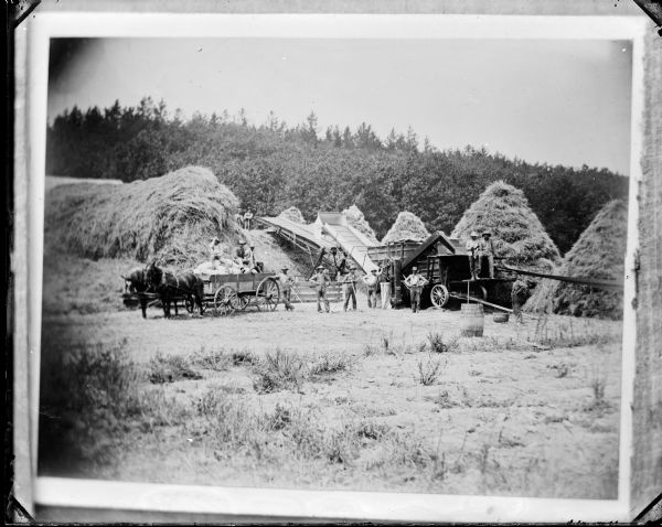A copy photograph of a threshing crew standing with wagon and threshing machine among cone-shaped haystacks.