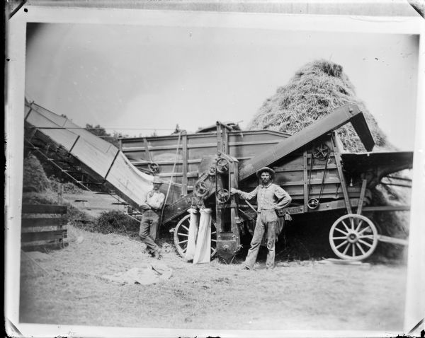 Copy photograph of two men posing next to a thresher. A haystack is in the background.