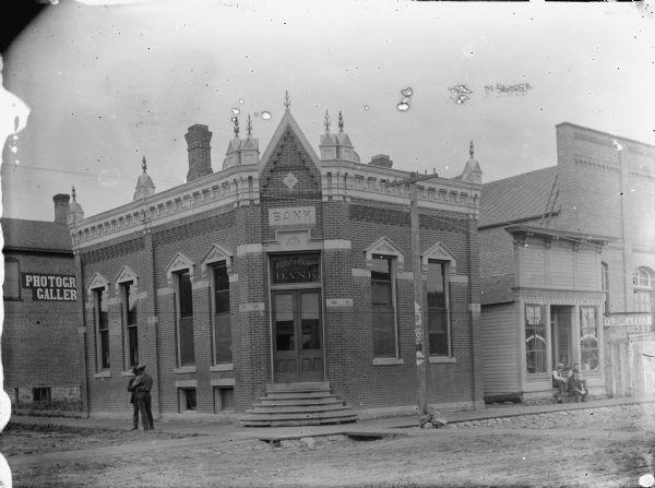 View across street towards the Jackson County Bank located at the southwest corner of First and Main Streets. Two men are standing on the sidewalk on the left. Two men are sitting in front of a storefront on the right.