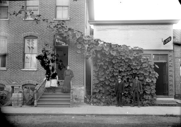 View from street towards the facades of Dr. Penton's Office, the telephone company and Van Schaick's photography studio. Two men are standing on the sidewalk. A woman and two girls are standing on the steps on the left. A vine is growing on a wall in front of the doctor's office, and along a wire above the telephone company entrance.