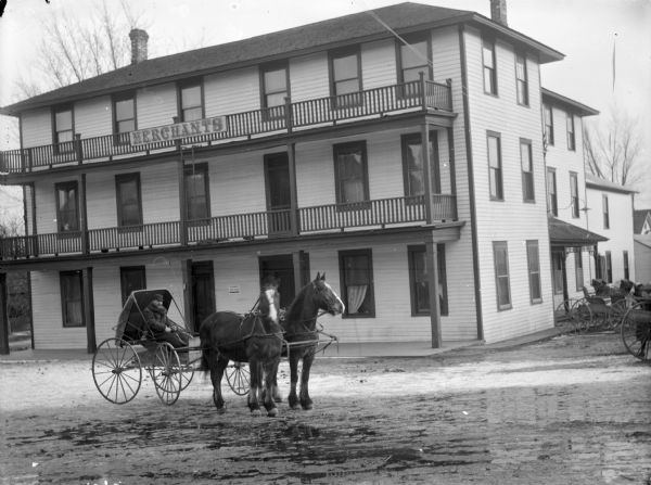View across street of Merchant's Inn located at the southwest corner of S. First and Fillmore Streets. A man in a buggy pulled by two horses is in the street in front. Two buggies are parked on the right.