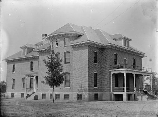 View across lawn towards a large brick house with two entrances. A man is sitting on the steps on the right side. A single tree is growing in the lawn. Identified as the Jackson County House built in 1905.